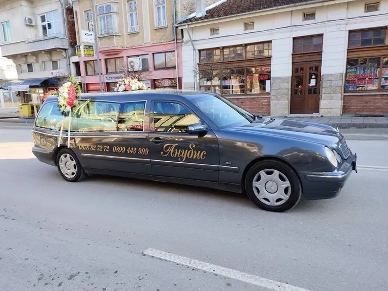 Сивата катафалка на агенция „Анубис“ с цветя на покрива